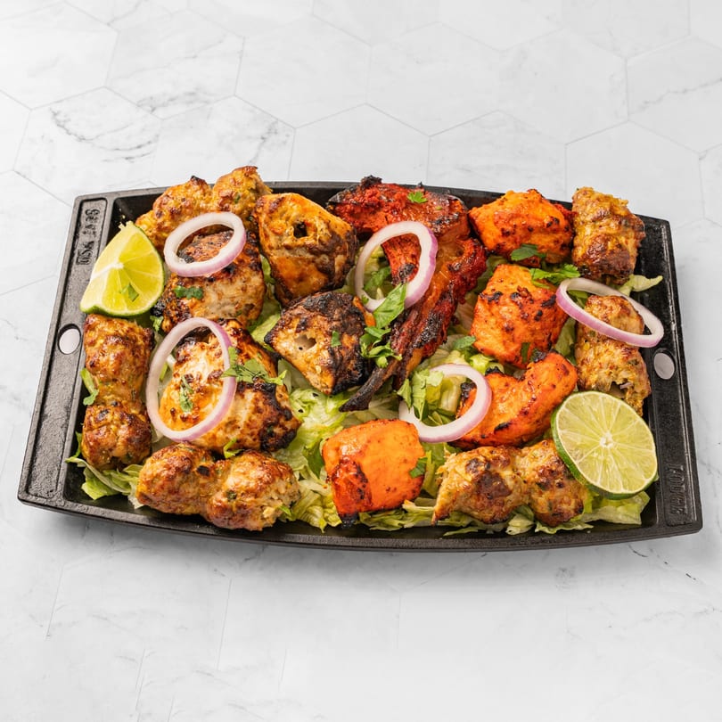 A black platter filled with various Indian kebabs, garnished with lime wedges and red onion rings.