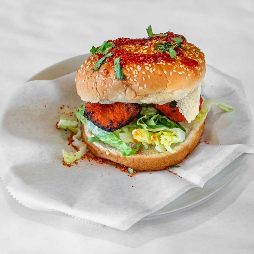 Indian chicken tikka burger with lettuce, red sauce, and cilantro on a sesame seed bun.