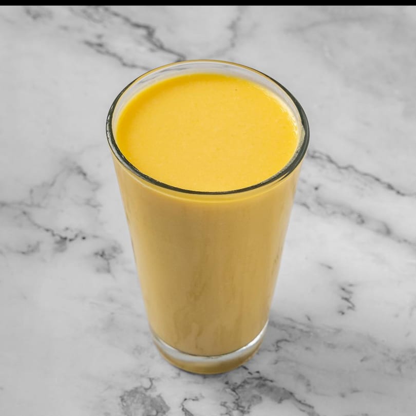 A tall glass of creamy yellow mango lassi sits on a white marble surface.