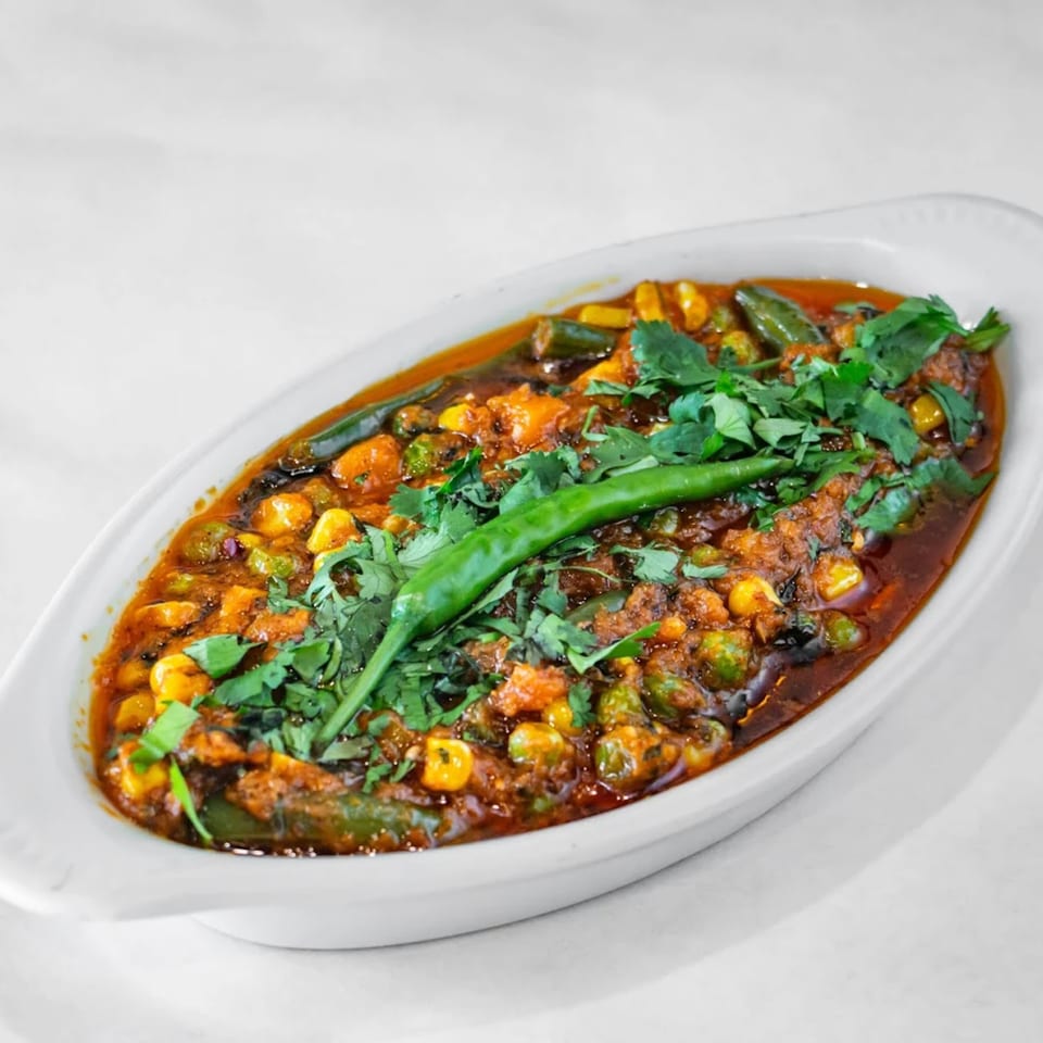 A white oval dish filled with a vibrant Indian vegetable curry, garnished with fresh cilantro and a green chili.