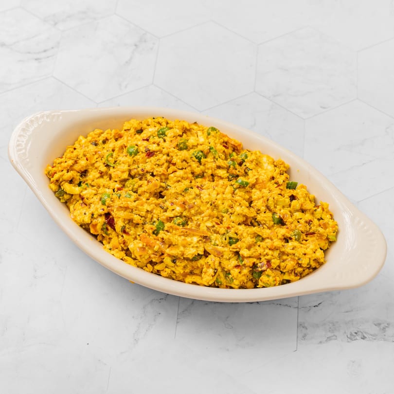 A beige oval dish filled with vibrant yellow scrambled paneer, green peas, and spices.
