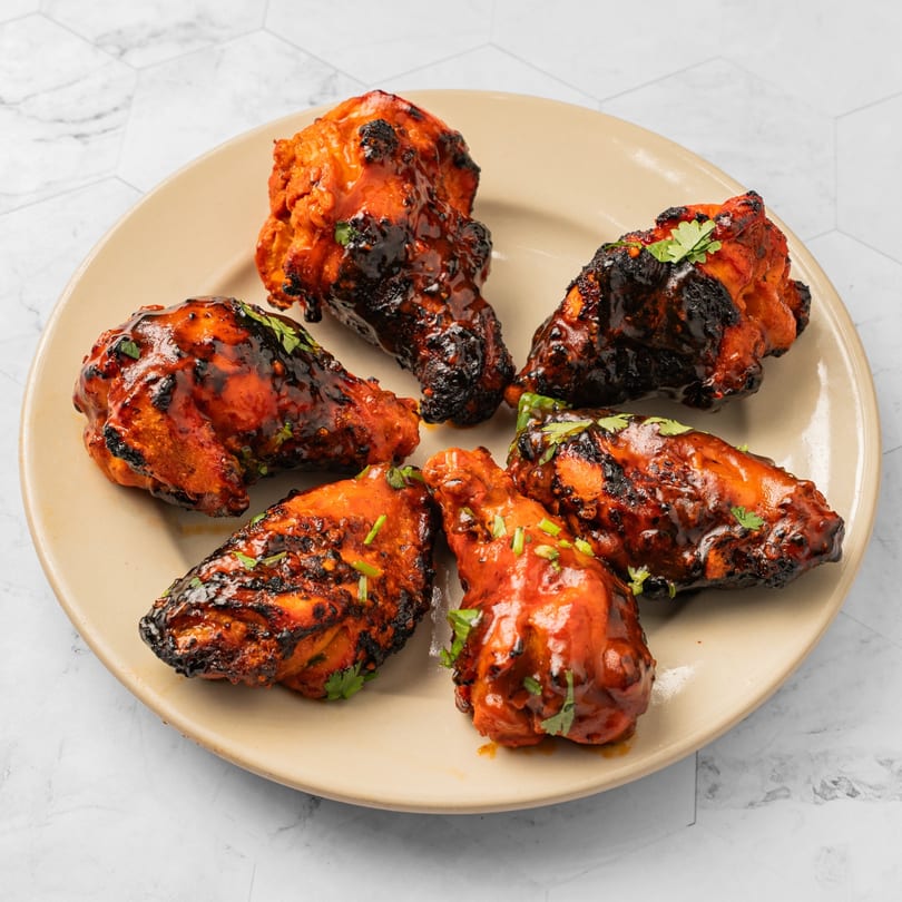 Six Indian-style grilled chicken drumsticks with red sauce and green herbs on a beige plate.