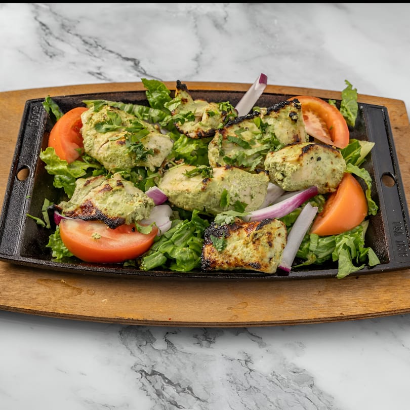 Indian chicken tikka with green marinade, lettuce, tomatoes, and red onions on a sizzling platter.