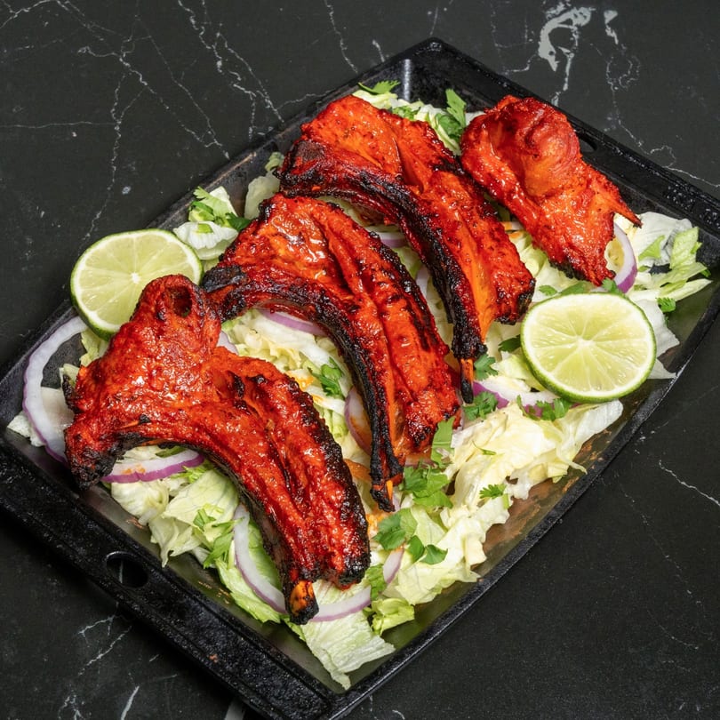 Four red tandoori lamb chops on a bed of lettuce, red onion, and lime slices.