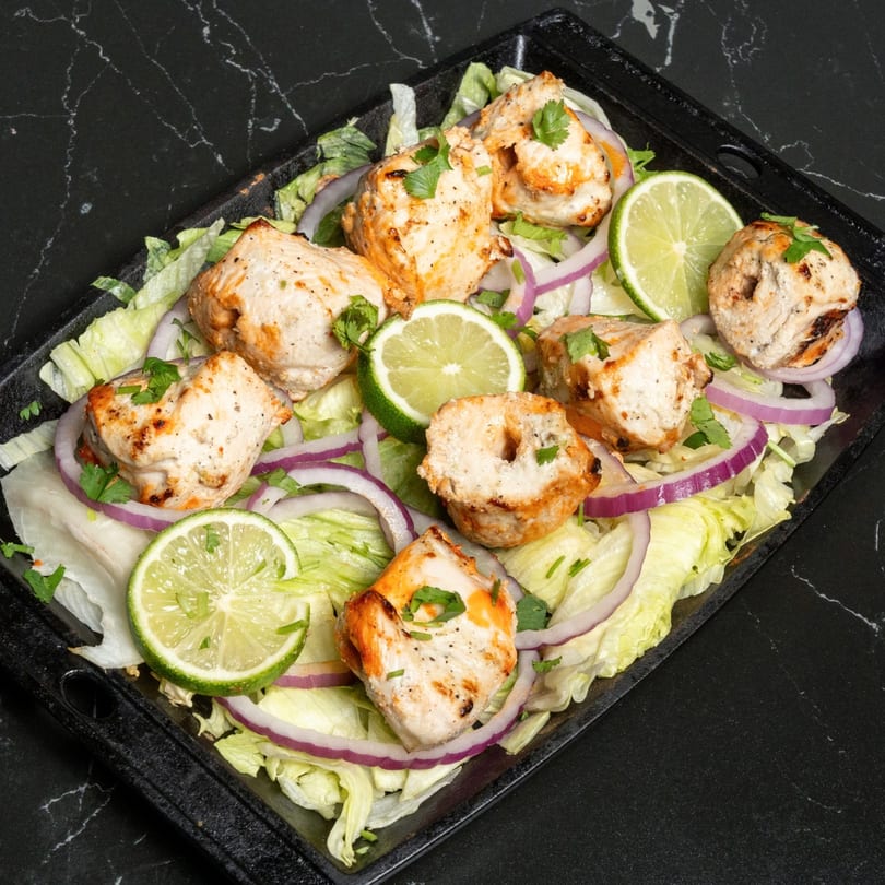 Chicken tikka pieces on a bed of lettuce and red onion, garnished with lime slices and cilantro.