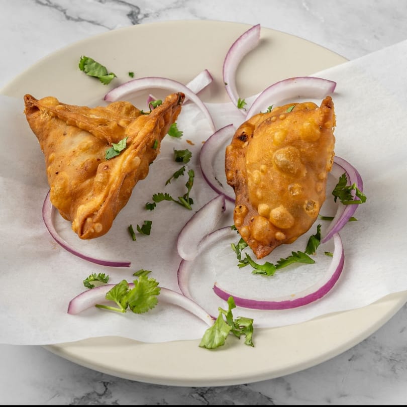 Two golden-brown Indian samosas garnished with red onion slices and fresh cilantro on a white plate.